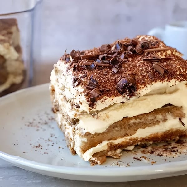 A slice of tiramisu with chocolate shavings on a white plate, with a coffee cup in the background.