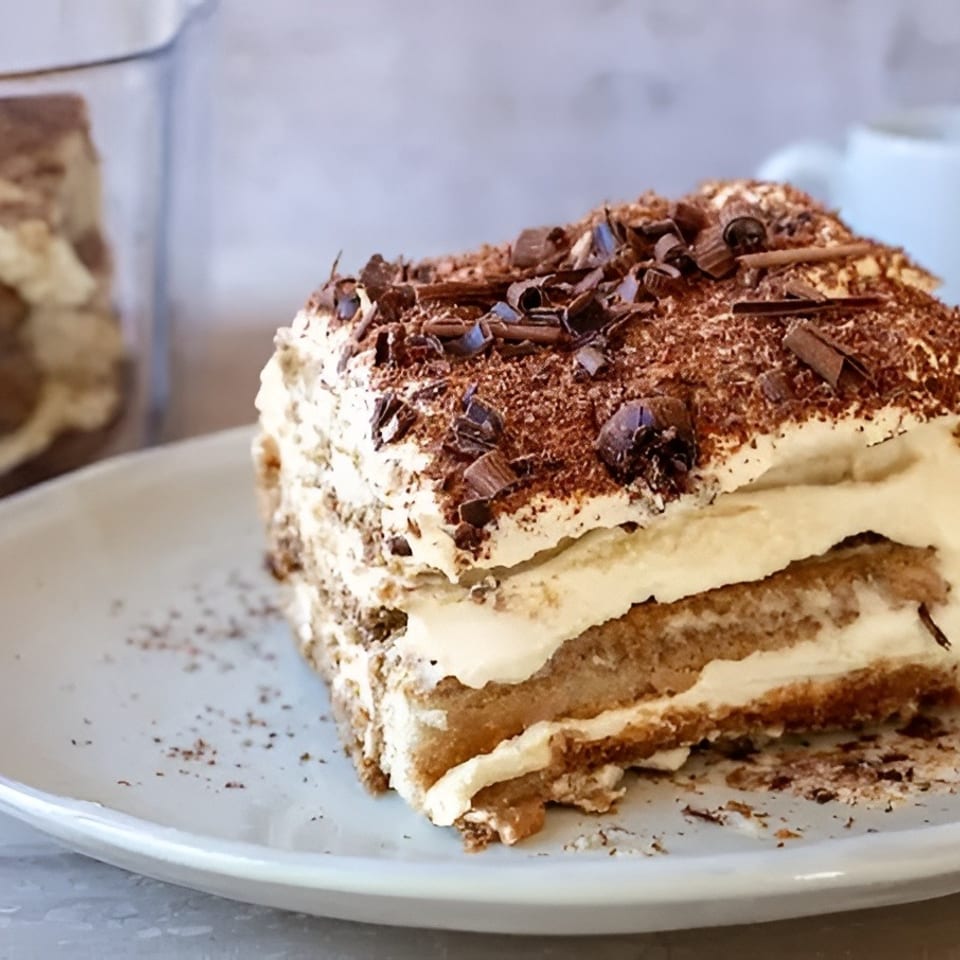 A slice of tiramisu with chocolate shavings on a white plate, with a coffee cup in the background.
