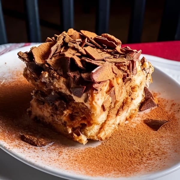 A slice of tiramisu with chocolate shavings and cocoa powder on a white plate.